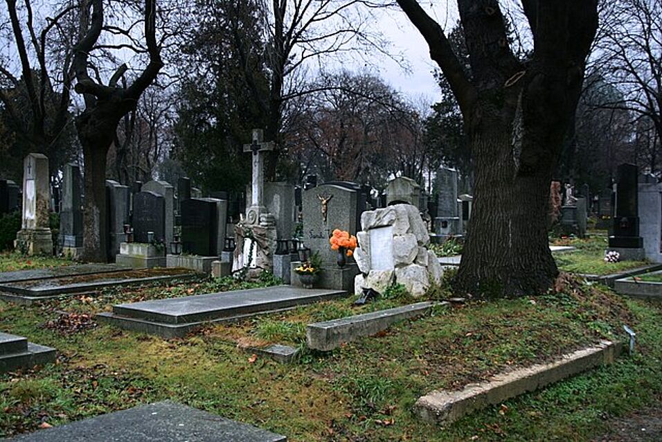 Friedhof während der Dämmerung, oranger Blumenschmuck auf einem Grab, kahle Bäume im Hintergrund