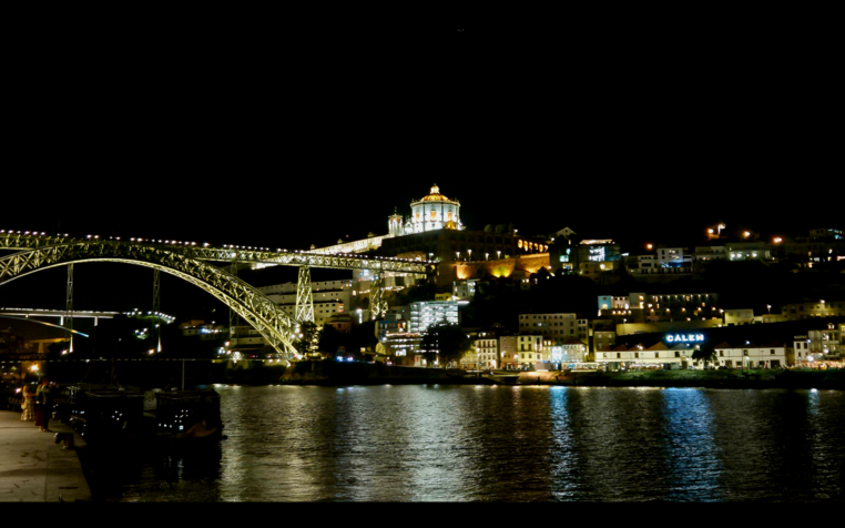 Die beleuchtete Altstadt Portos am Fluss Douro bei Nacht; in der Mitte des Bildes ist eine große Brücke zu sehen; die Lichter spiegeln sich im Wasser