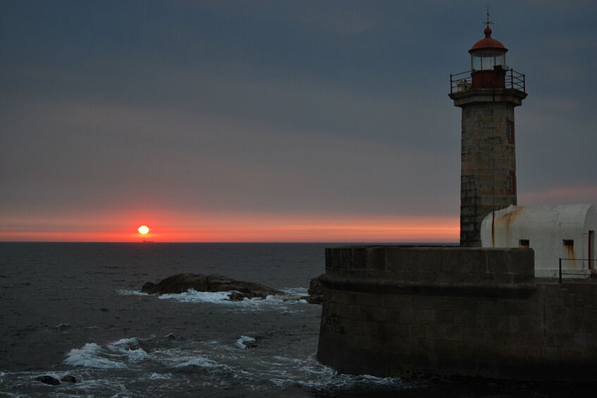 Sonnenuntergang am Meer; im Vordergrund ist ein Leuchtturm im Hafen zu sehen